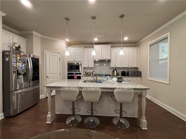 kitchen featuring appliances with stainless steel finishes, decorative light fixtures, white cabinetry, an island with sink, and light stone countertops