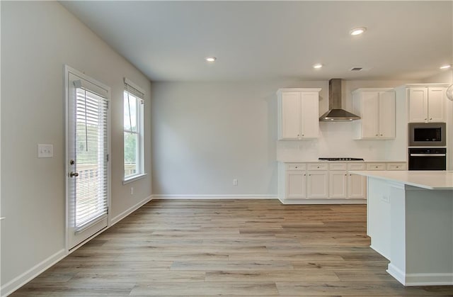 kitchen with stainless steel microwave, black oven, white cabinetry, light countertops, and wall chimney exhaust hood