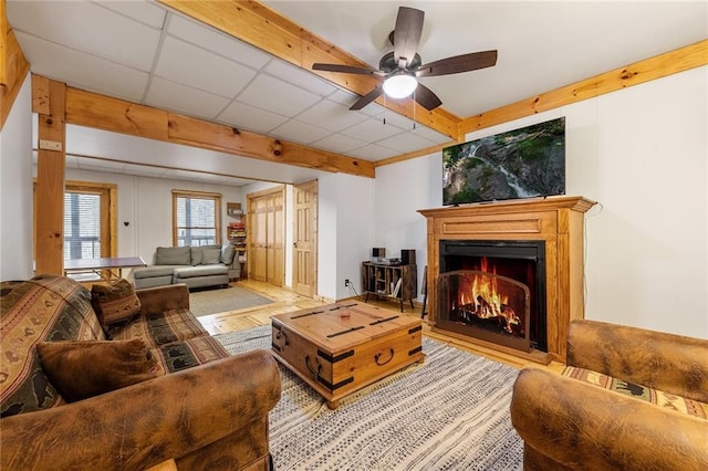 living area featuring beam ceiling, wood finished floors, a lit fireplace, a paneled ceiling, and ceiling fan