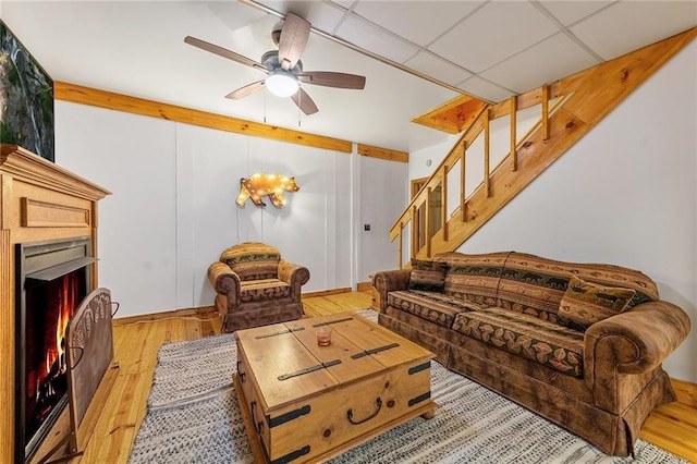 living room featuring stairway, a ceiling fan, wood finished floors, a warm lit fireplace, and a paneled ceiling