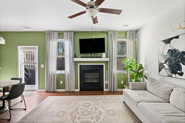 living room featuring visible vents, a glass covered fireplace, wood finished floors, baseboards, and ceiling fan