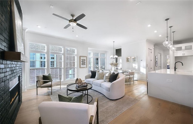 living room with light hardwood / wood-style flooring, a brick fireplace, crown molding, ceiling fan with notable chandelier, and sink