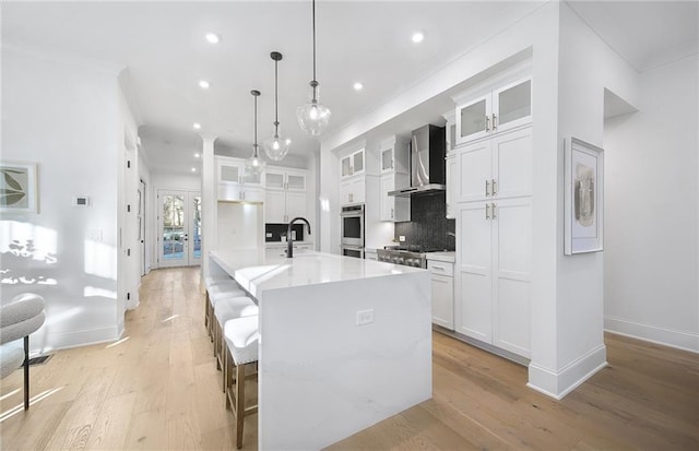 kitchen with decorative light fixtures, white cabinets, an island with sink, decorative backsplash, and wall chimney range hood