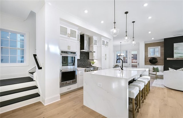 kitchen with a center island with sink, pendant lighting, wall chimney exhaust hood, white cabinetry, and backsplash