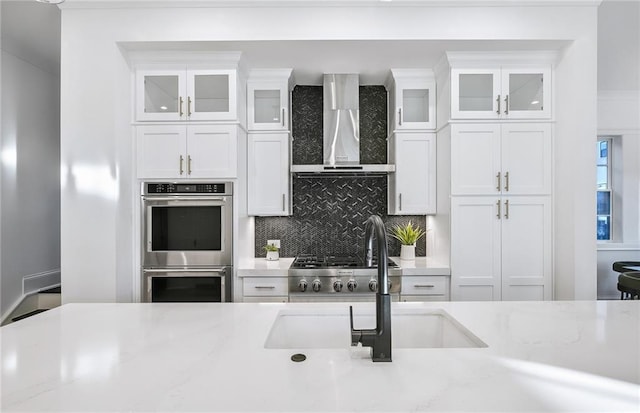 kitchen featuring wall chimney exhaust hood, decorative backsplash, white cabinetry, double oven, and sink