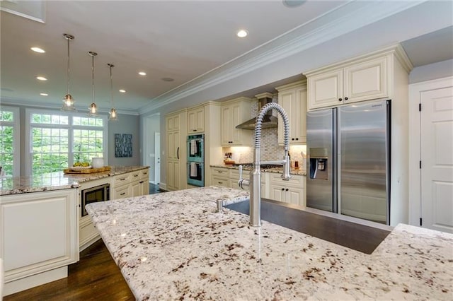 kitchen featuring light stone countertops, hanging light fixtures, stainless steel appliances, cream cabinetry, and ornamental molding