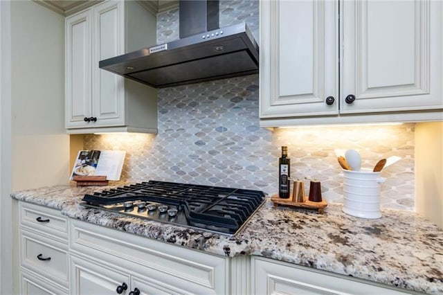 kitchen with stainless steel gas stovetop, backsplash, white cabinets, wall chimney range hood, and light stone counters