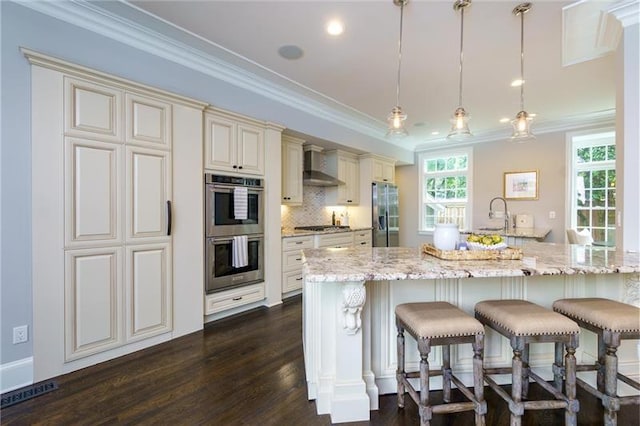 kitchen with wall chimney exhaust hood, hanging light fixtures, light stone counters, backsplash, and appliances with stainless steel finishes