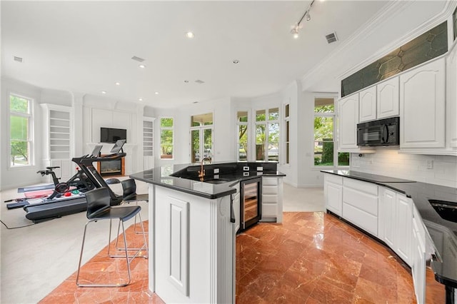 kitchen featuring an island with sink, beverage cooler, white cabinetry, decorative backsplash, and ornamental molding