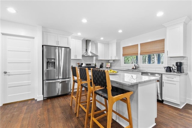 kitchen featuring a kitchen island, a sink, white cabinets, appliances with stainless steel finishes, and wall chimney range hood