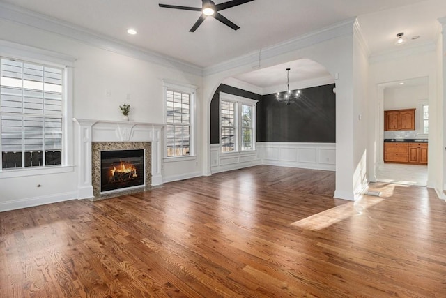 unfurnished living room featuring a premium fireplace, a wainscoted wall, ceiling fan with notable chandelier, and wood finished floors