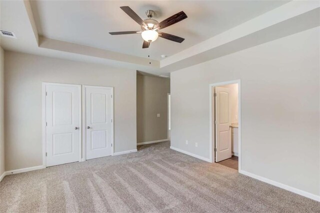 unfurnished bedroom with a raised ceiling, ensuite bath, ceiling fan, and light colored carpet