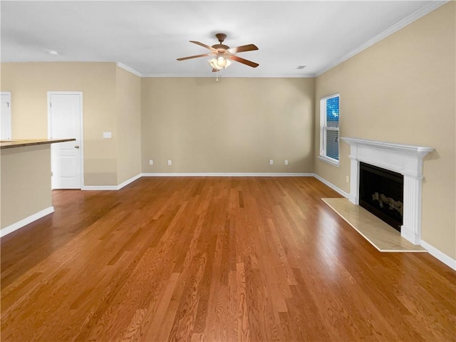 unfurnished living room featuring a fireplace with raised hearth, wood finished floors, a ceiling fan, baseboards, and crown molding