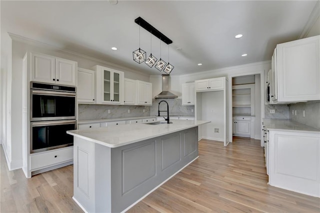 kitchen featuring wall chimney range hood, stainless steel double oven, a sink, light wood-style floors, and backsplash