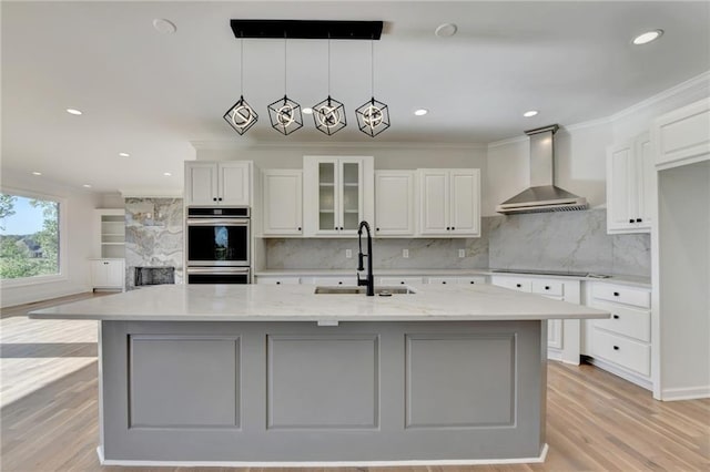 kitchen featuring wall chimney range hood, a sink, light wood finished floors, double oven, and tasteful backsplash