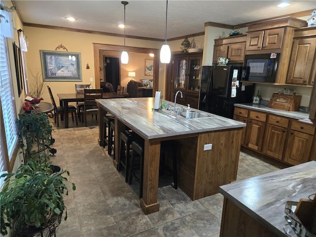 kitchen with crown molding, brown cabinetry, a kitchen island with sink, a sink, and black appliances