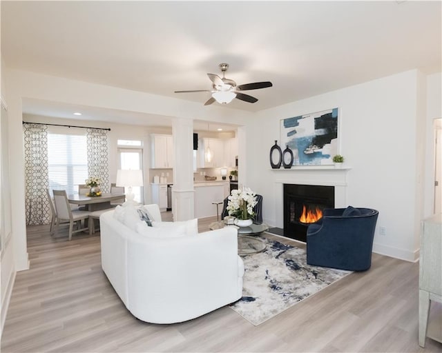 living room with ceiling fan, a lit fireplace, baseboards, and light wood-style flooring