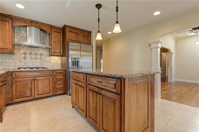 kitchen with arched walkways, appliances with stainless steel finishes, wall chimney exhaust hood, brown cabinetry, and ornate columns