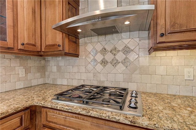 kitchen featuring backsplash, glass insert cabinets, stainless steel gas cooktop, exhaust hood, and brown cabinetry