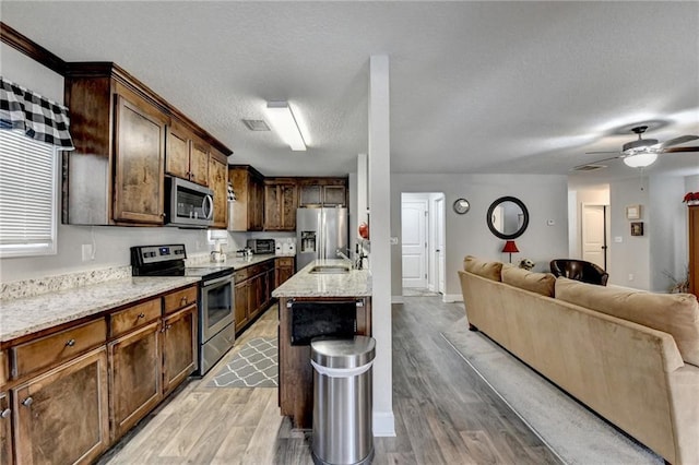 kitchen with a center island with sink, light wood-type flooring, dark brown cabinets, light stone counters, and stainless steel appliances