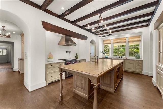 kitchen featuring wall chimney range hood, beamed ceiling, stainless steel stove, a chandelier, and a center island with sink