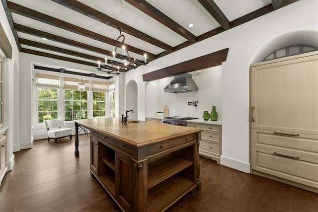 kitchen featuring wall chimney exhaust hood, beamed ceiling, sink, a kitchen island with sink, and cream cabinetry