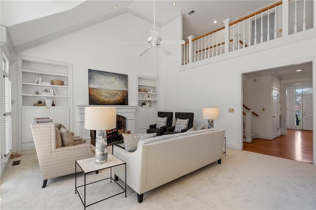 carpeted living area with built in shelves, stairs, a fireplace, high vaulted ceiling, and a ceiling fan