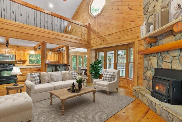 living room with wood walls, a wood stove, high vaulted ceiling, light hardwood / wood-style floors, and a stone fireplace