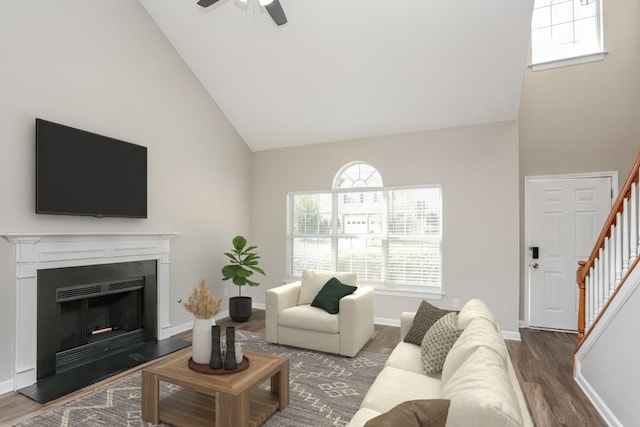 living area featuring high vaulted ceiling, stairs, wood finished floors, a ceiling fan, and a fireplace with raised hearth