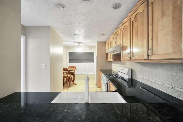 kitchen with gas range gas stove, under cabinet range hood, a textured ceiling, ceiling fan, and dark stone counters
