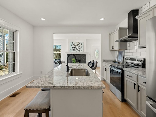 kitchen featuring gray cabinetry, wall chimney range hood, sink, stainless steel electric range oven, and an island with sink