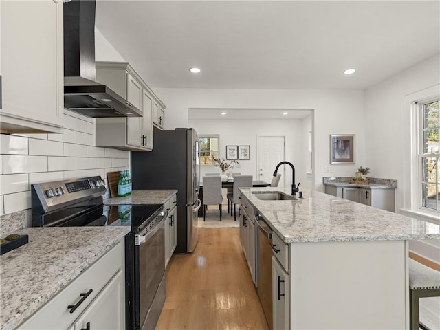 kitchen featuring sink, wall chimney exhaust hood, an island with sink, light hardwood / wood-style floors, and appliances with stainless steel finishes