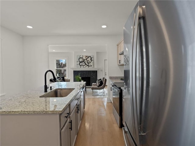 kitchen featuring sink, a high end fireplace, an island with sink, appliances with stainless steel finishes, and light wood-type flooring