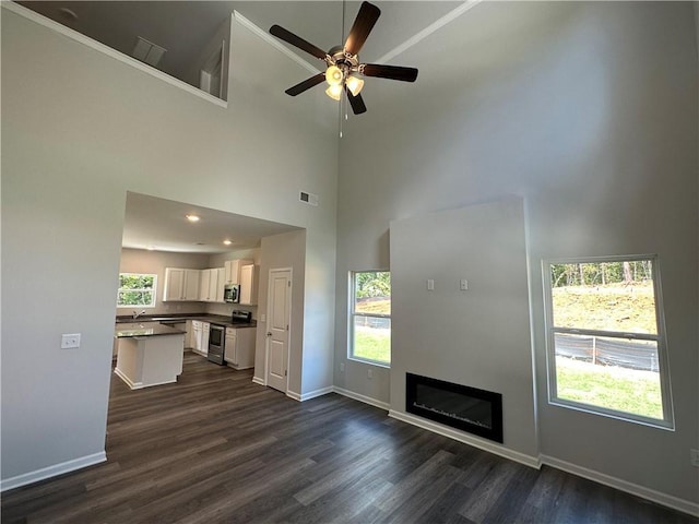 unfurnished living room with a high ceiling, ceiling fan, dark wood-style floors, baseboards, and a glass covered fireplace