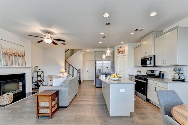 kitchen with backsplash, a center island with sink, light wood-type flooring, appliances with stainless steel finishes, and decorative light fixtures