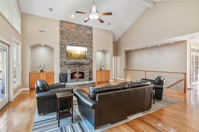 living room featuring light wood-type flooring, ceiling fan, high vaulted ceiling, beamed ceiling, and a stone fireplace