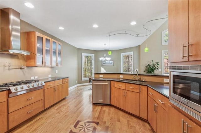 kitchen featuring wall chimney range hood, sink, light wood-type flooring, appliances with stainless steel finishes, and decorative light fixtures