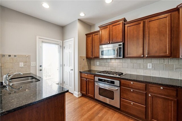 kitchen featuring light wood finished floors, dark stone counters, brown cabinetry, appliances with stainless steel finishes, and a sink