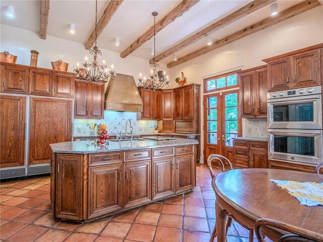 kitchen with custom exhaust hood, a notable chandelier, sink, double oven, and a kitchen island with sink