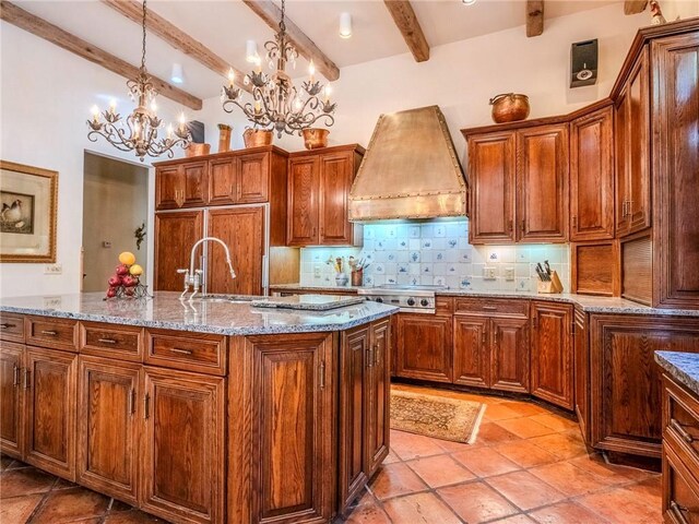 kitchen featuring beamed ceiling, pendant lighting, a notable chandelier, premium range hood, and an island with sink