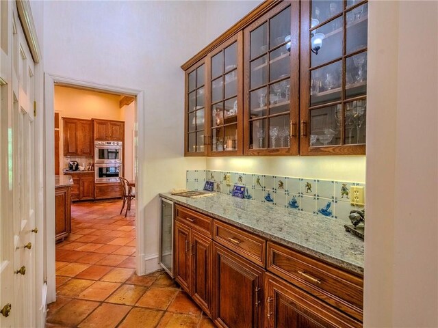 bar featuring light tile patterned floors, stainless steel double oven, light stone counters, wine cooler, and decorative backsplash