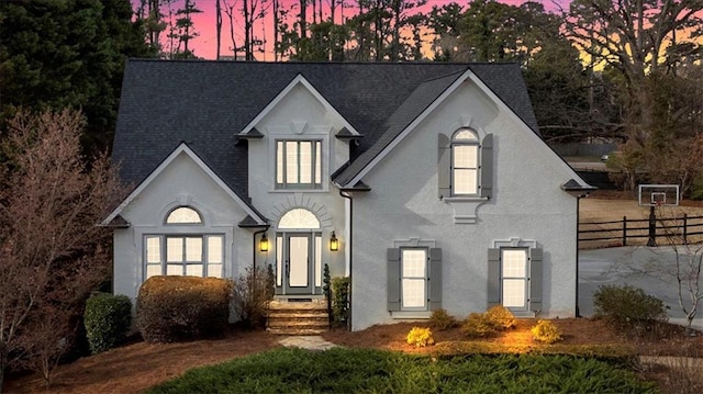 french provincial home featuring a shingled roof, fence, and stucco siding