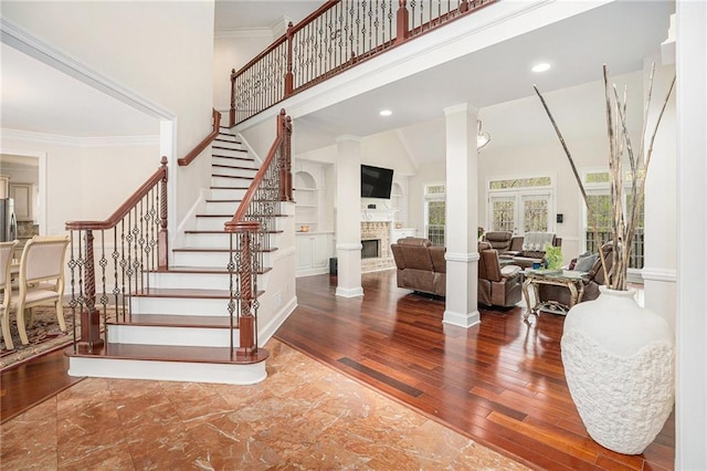 staircase featuring a high ceiling, a brick fireplace, crown molding, ornate columns, and wood finished floors