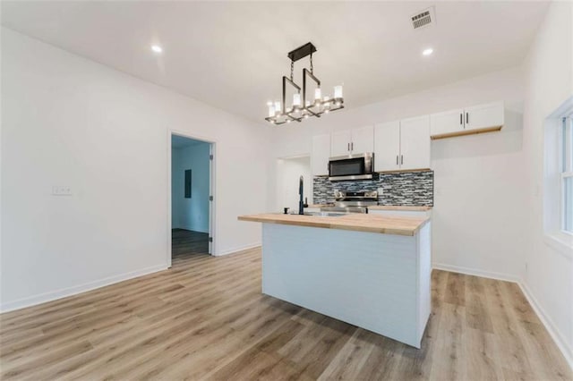 kitchen featuring appliances with stainless steel finishes, a center island with sink, white cabinetry, hanging light fixtures, and butcher block counters