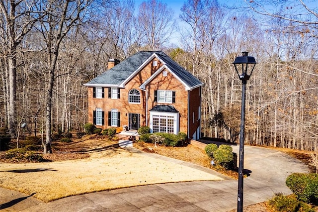 view of front of property with driveway, brick siding, a chimney, and a view of trees