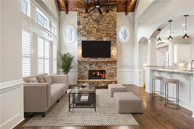 living area featuring beamed ceiling, a stone fireplace, wainscoting, wooden ceiling, and dark wood-style flooring