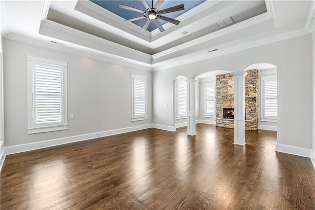 empty room featuring ceiling fan, a tray ceiling, a stone fireplace, ornate columns, and dark wood-style flooring