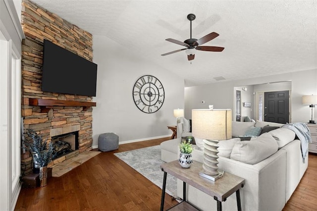 living room featuring ceiling fan, wood finished floors, vaulted ceiling, a textured ceiling, and a stone fireplace