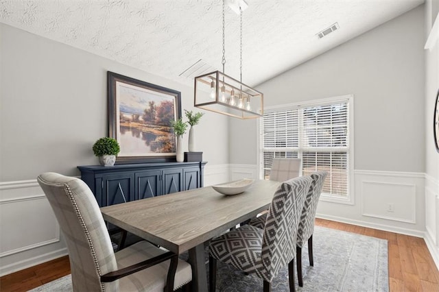 dining area with lofted ceiling, a textured ceiling, light wood-style flooring, and a wainscoted wall