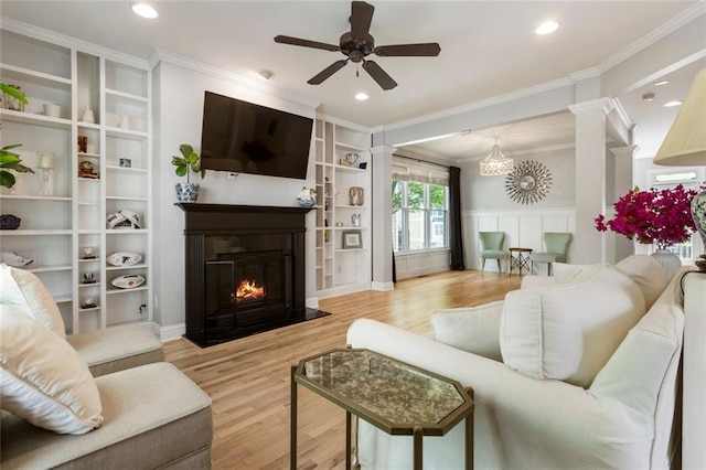 living area with a ceiling fan, decorative columns, crown molding, a fireplace with flush hearth, and wood finished floors